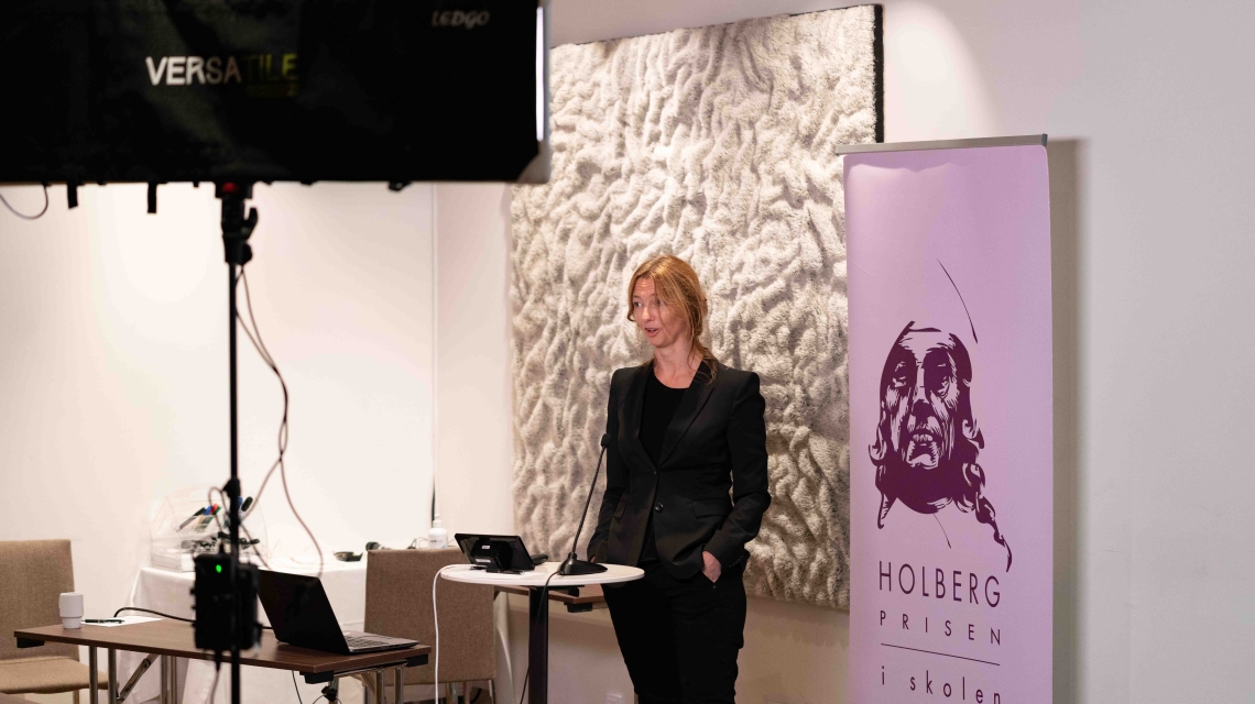 Bilde av fagkonsulent Marie von der Lippe stående i oppsettet for digital avvikling: Foran en roll up med Holbergprisen i skolens logo, og et bord med mikrofon, og bak en lyskaster. Foto: Thor Brødreskift